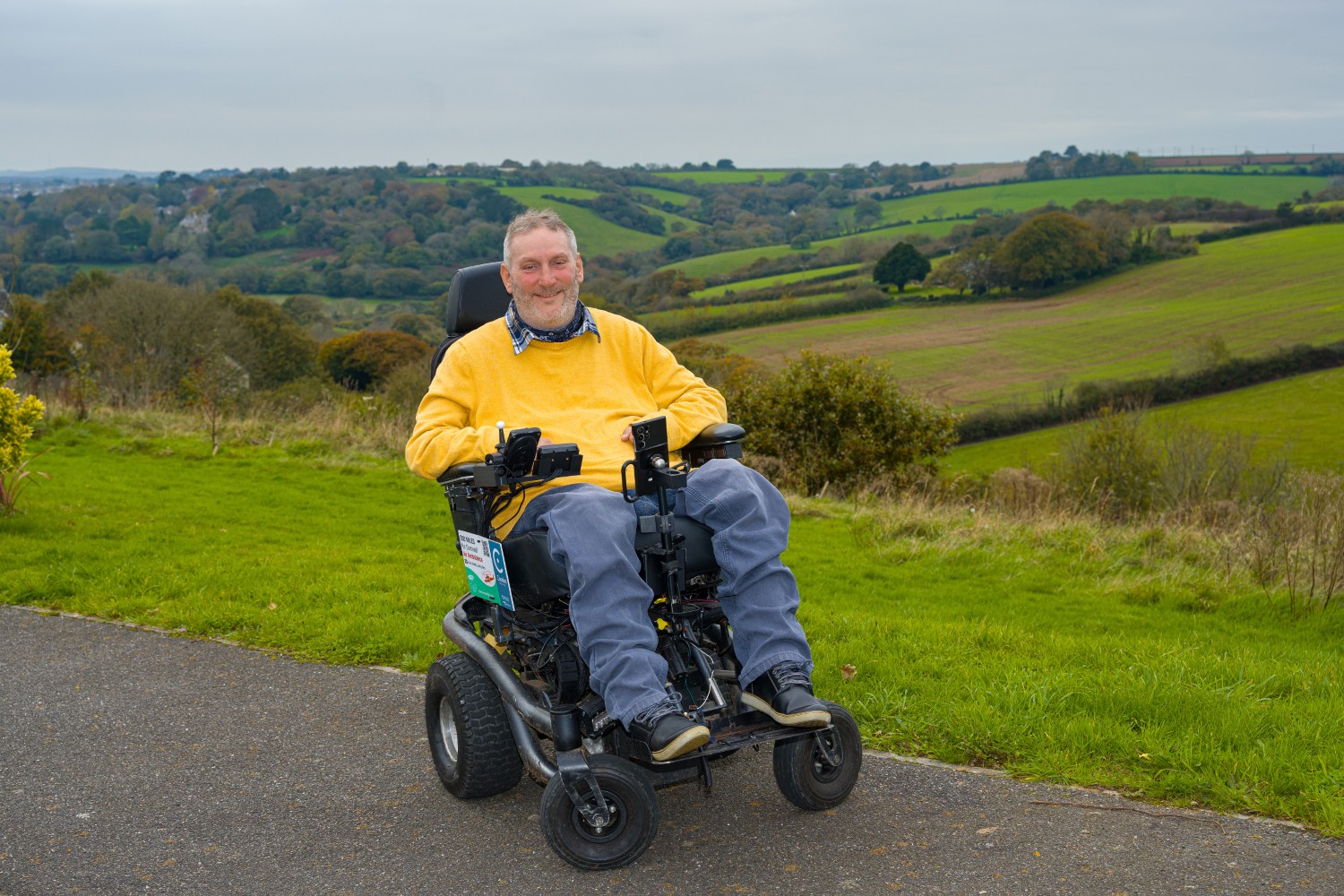 Steven Webb smiling in his electric wheelchair, outdoors with fields behind him